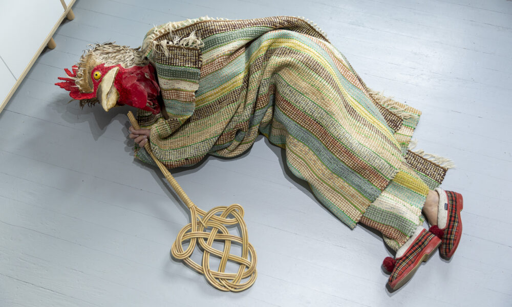 The Chicken wrapped in a carpet on the floor with a carpet beater in hand.