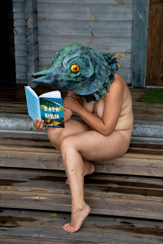 A naked person in a Fish-head mask sitting on wet wooden steps mext to a summer cottage holding a book the cover of which reads "KALA KIRJA", "FISH BOOK" in Finnish.
